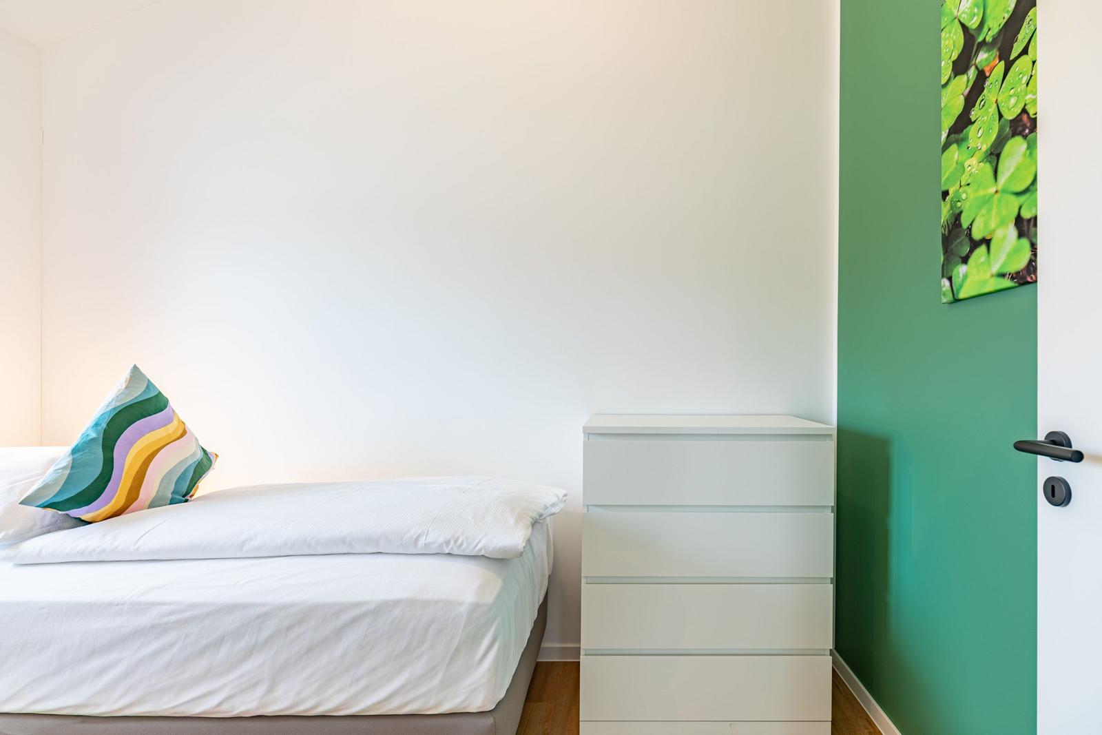 Schlafzimmer mit weißem Bett, Schrank und grüner Wand. Kissen mit buntem Muster.