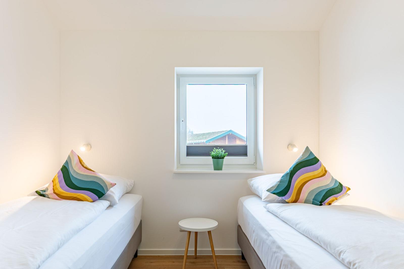 Zwei Einzelbetten mit bunten Kissen stehen gegenüber einem Fenster mit Pflanze.