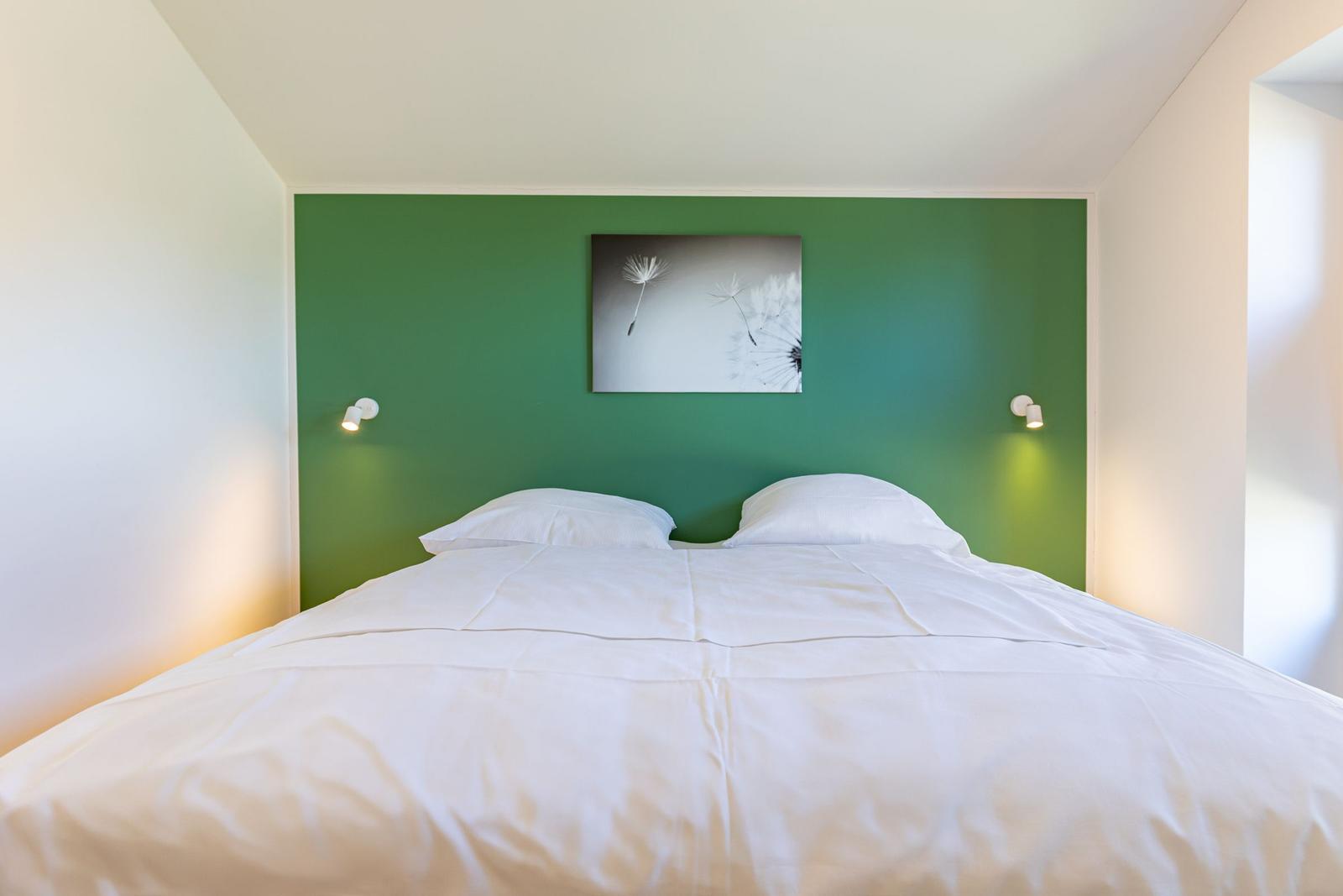 Schlafzimmer mit grünem Akzentwand, weißem Bett und Wandbild mit Löwenzahn.