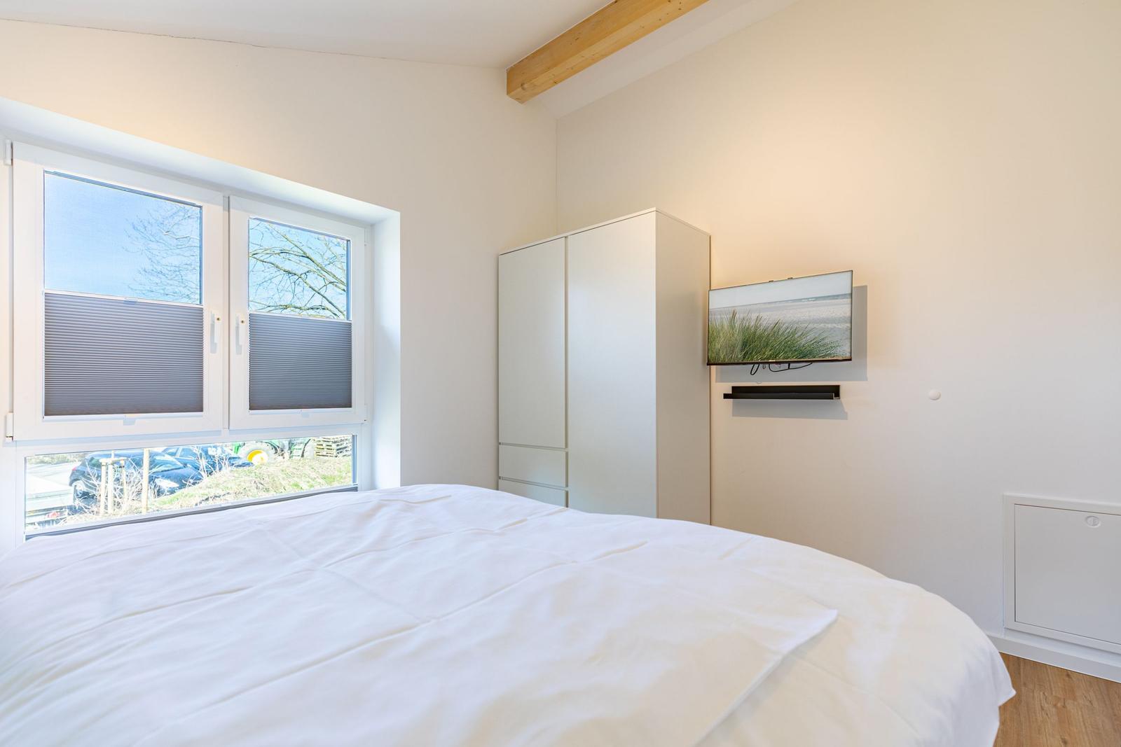 Schlafzimmer mit weißem Bett, Schrank und Wandbild. Fenster mit Jalousien und Holzbalken.