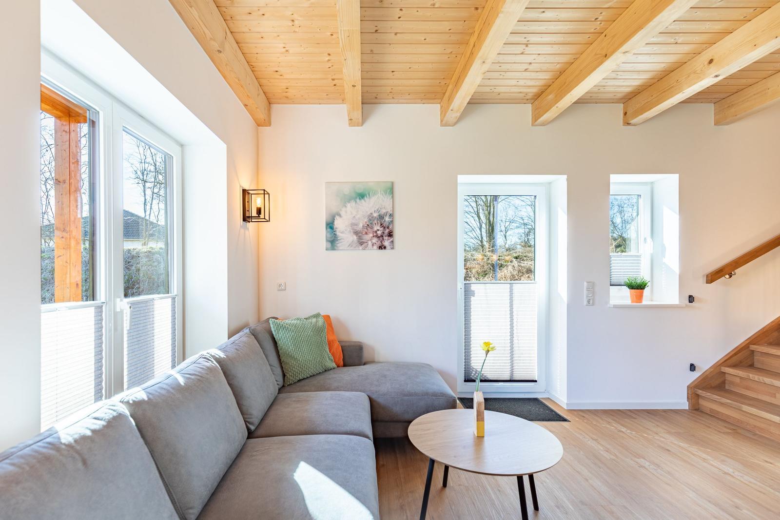 Wohnzimmer mit Ecksofa, Holzdecke und Fenstern. Tisch und Wandbild sind sichtbar.