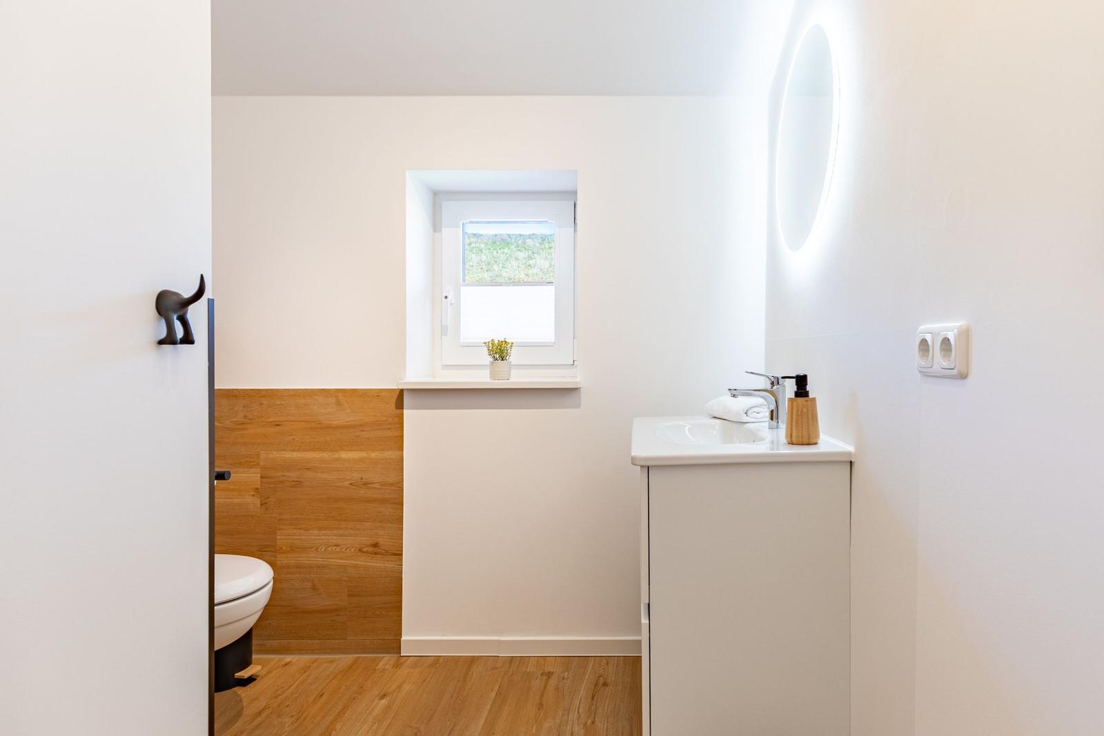 Badezimmer mit Waschbecken, Spiegel, Toilette und Fenster mit Blick auf Grün.