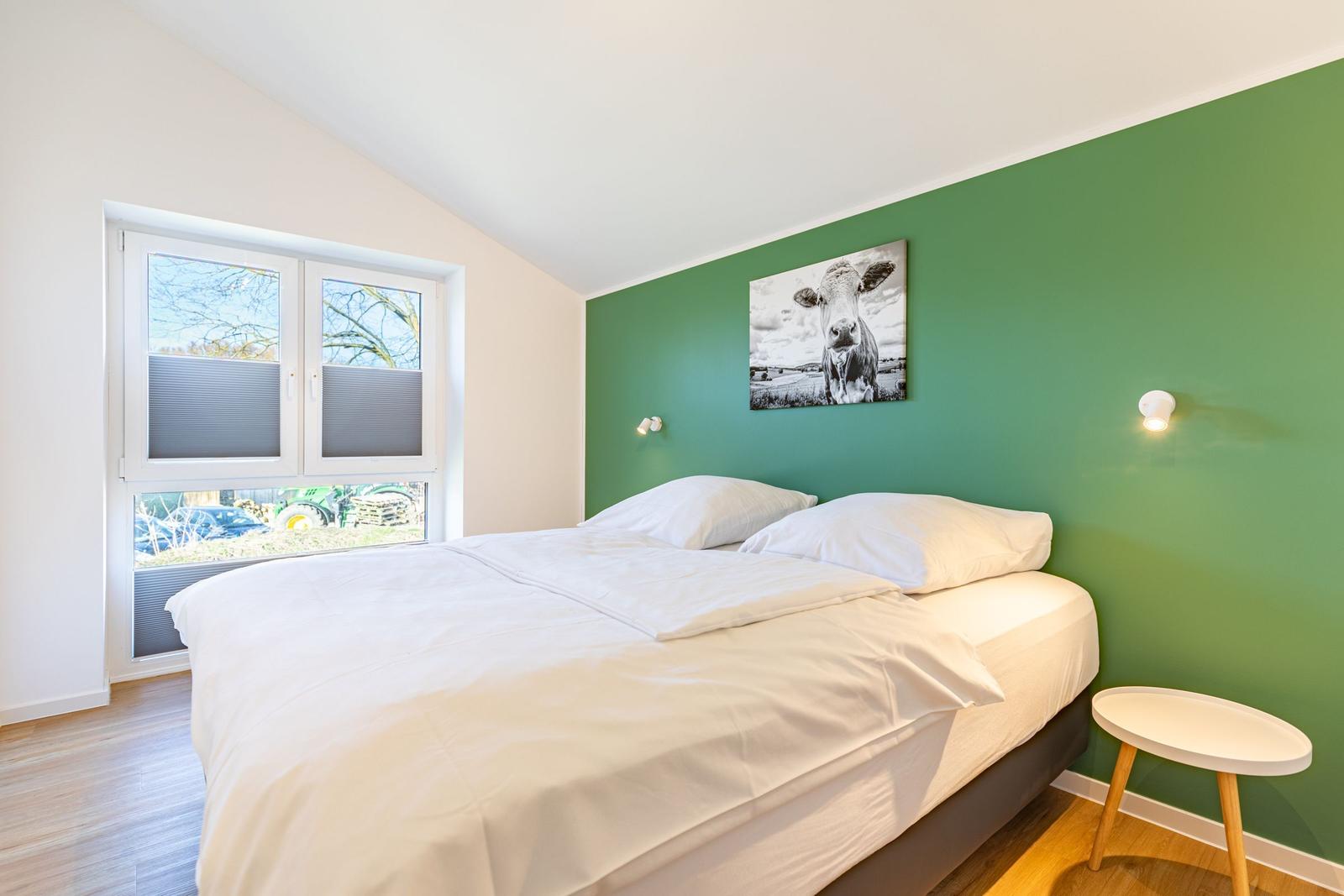 Schlafzimmer mit grünem Wandbereich, Bett und Fenster mit Blick auf Außenbereich.