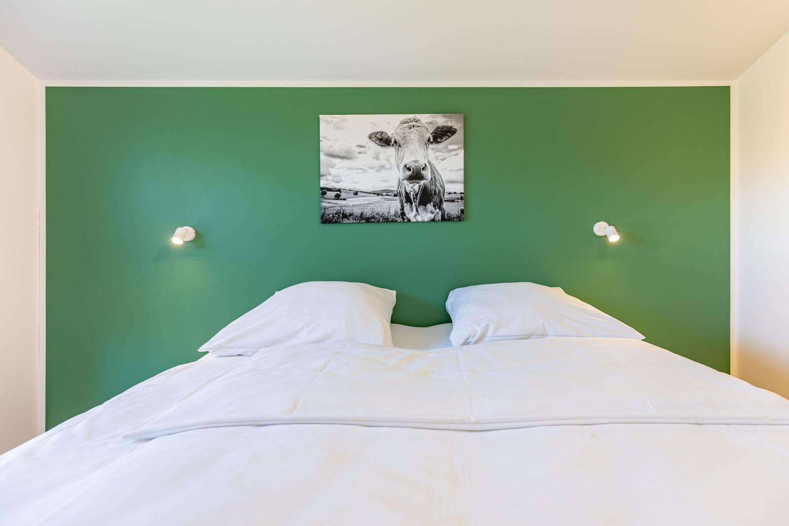 Schlafzimmer mit grünem Wandbild, Bett mit weißen Kissen und Decke.