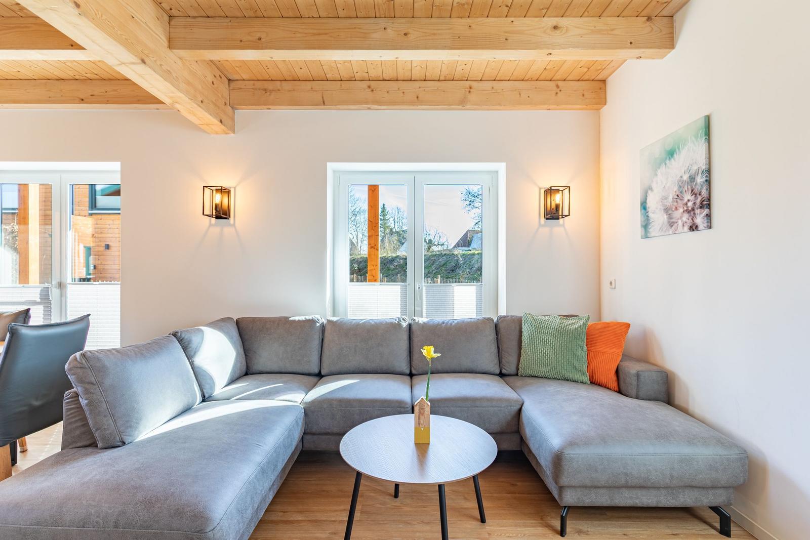 Ein Wohnzimmer mit grauem Sofa, Holzboden und Holzdeckenbalken. An der Wand hängt ein Bild.