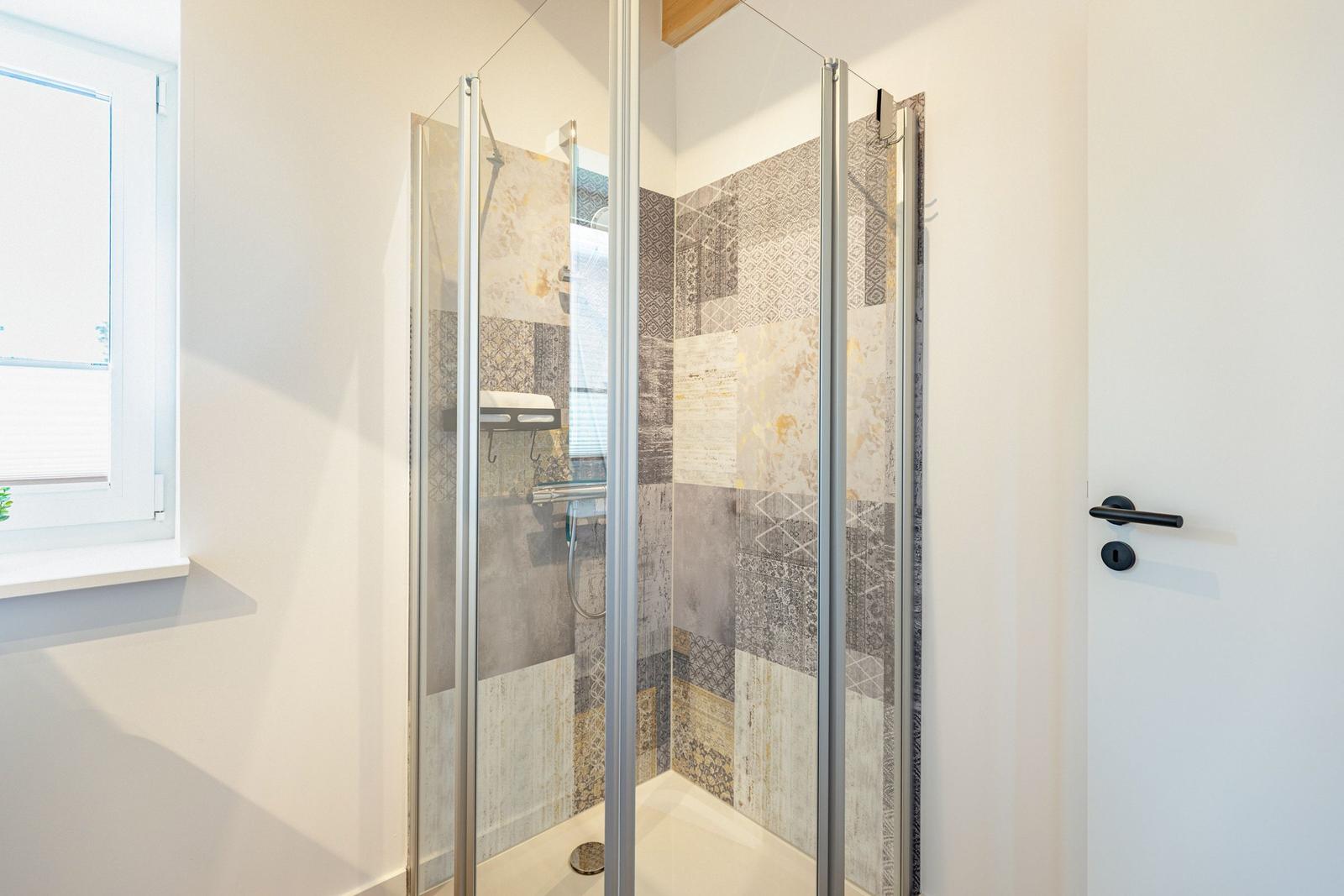 Dusche mit Glaswand und gemusterten Fliesen. Fenster und Tür sind sichtbar.
