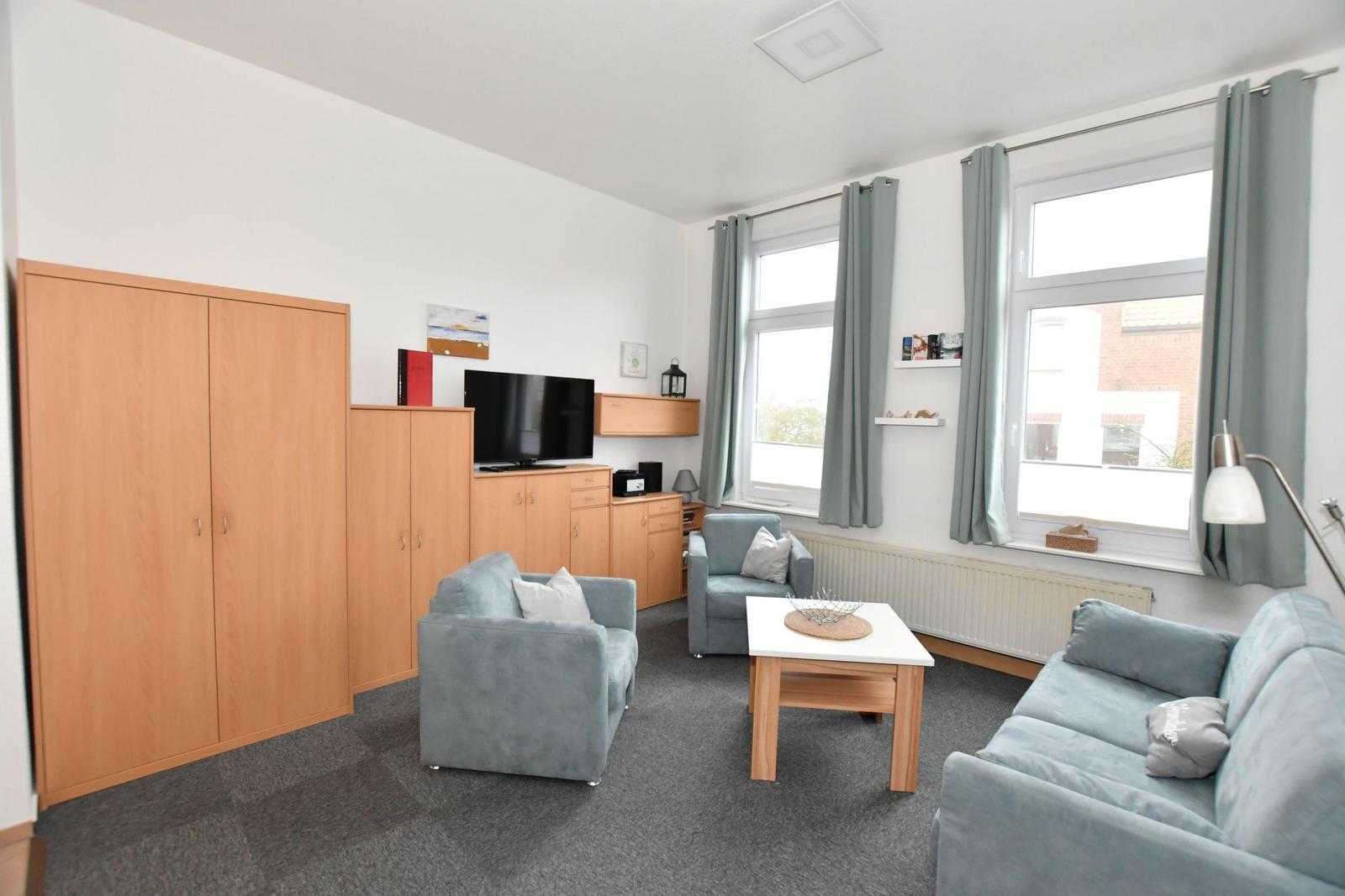 Wohnzimmer mit Sofa, Sesseln, TV und Fenstern mit Vorhängen.