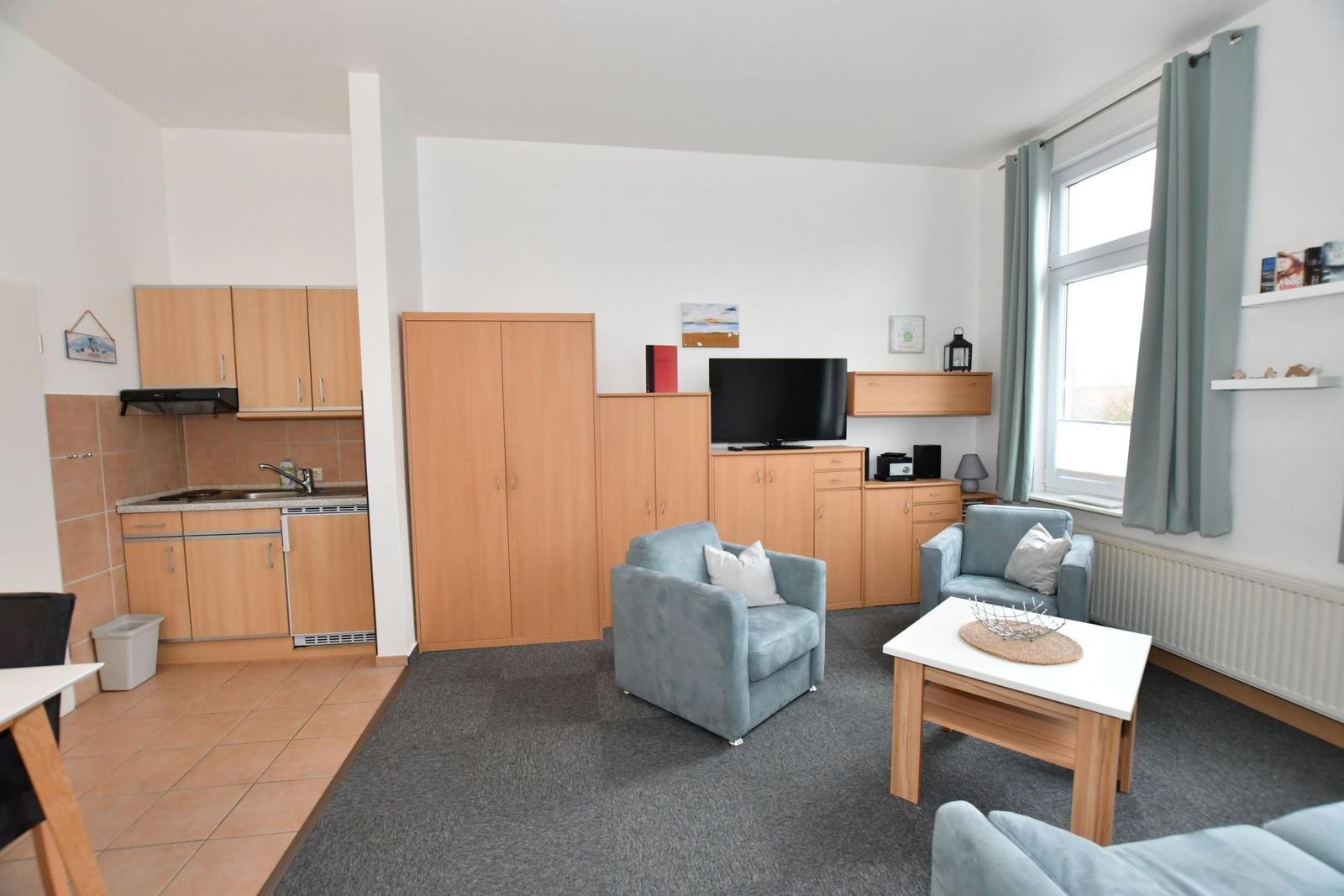 Wohnung mit Küche, Wohnzimmer, zwei Sesseln und Fernseher. Großer Fensterbereich mit Vorhängen.