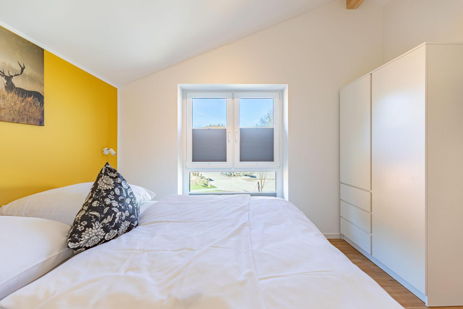 Schlafzimmer mit Bett, Fenster und Kleiderschrank. Gelbe Wand mit Bild.