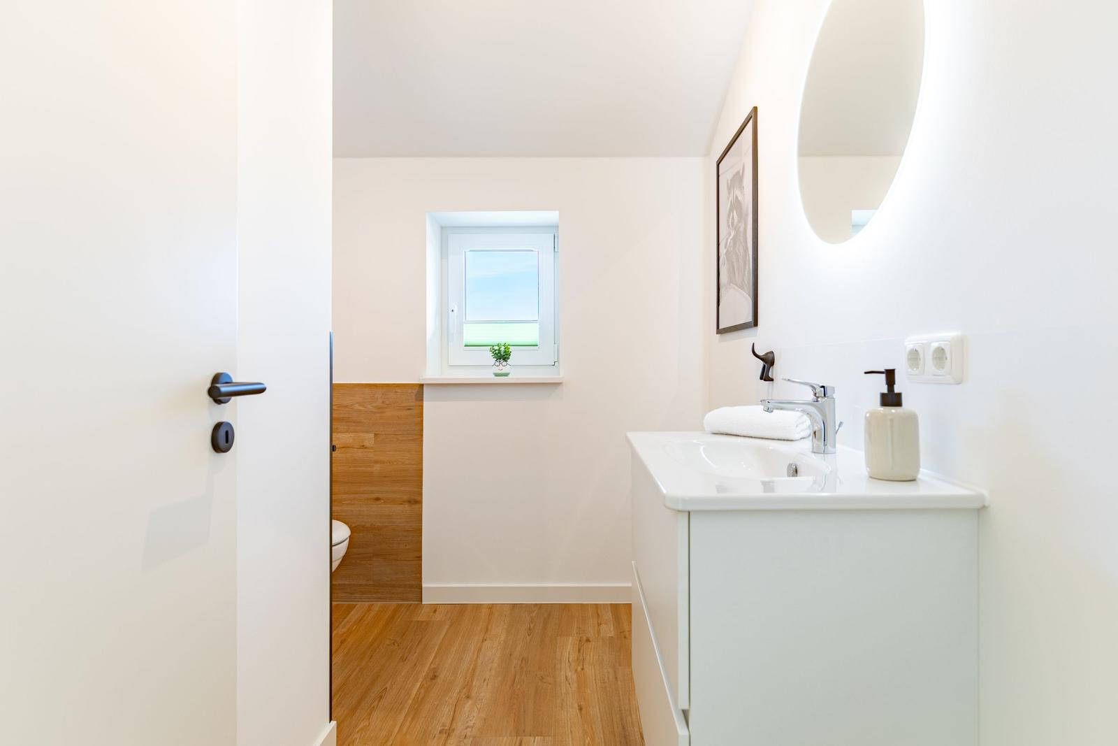 Badezimmer mit Waschtisch, Spiegel und Fenster. Holzfußboden und Toilette im Hintergrund.