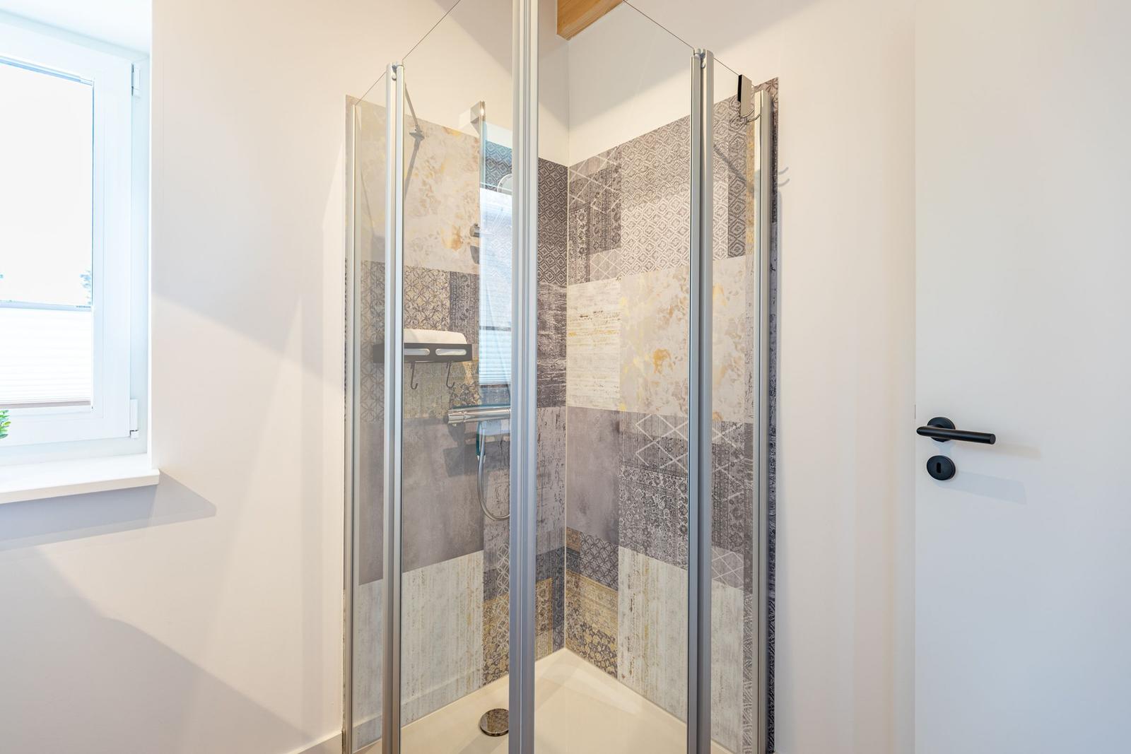 Dusche mit Glaswand und gemusterten Fliesen. Fenster und Tür sind sichtbar.