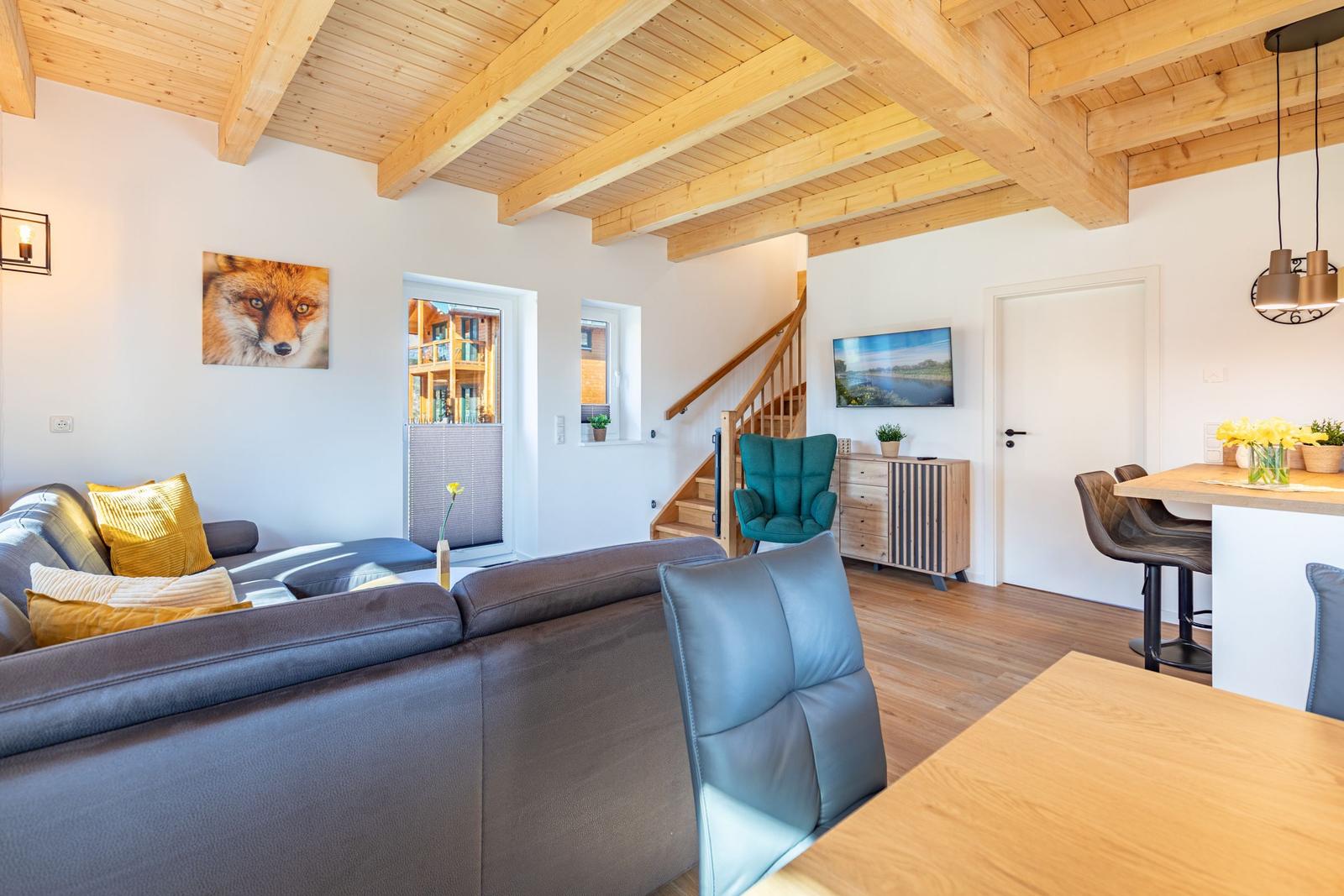 Wohn- und Essbereich mit Holzdecke, Sofa, TV und Tisch. Treppenhaus im Hintergrund.