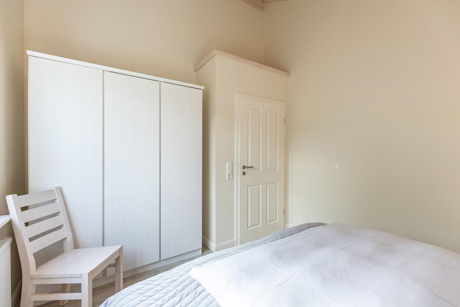 Schlafzimmer mit Bett, Kleiderschrank und Stuhl. Tür und Wandfarbe sind hell.