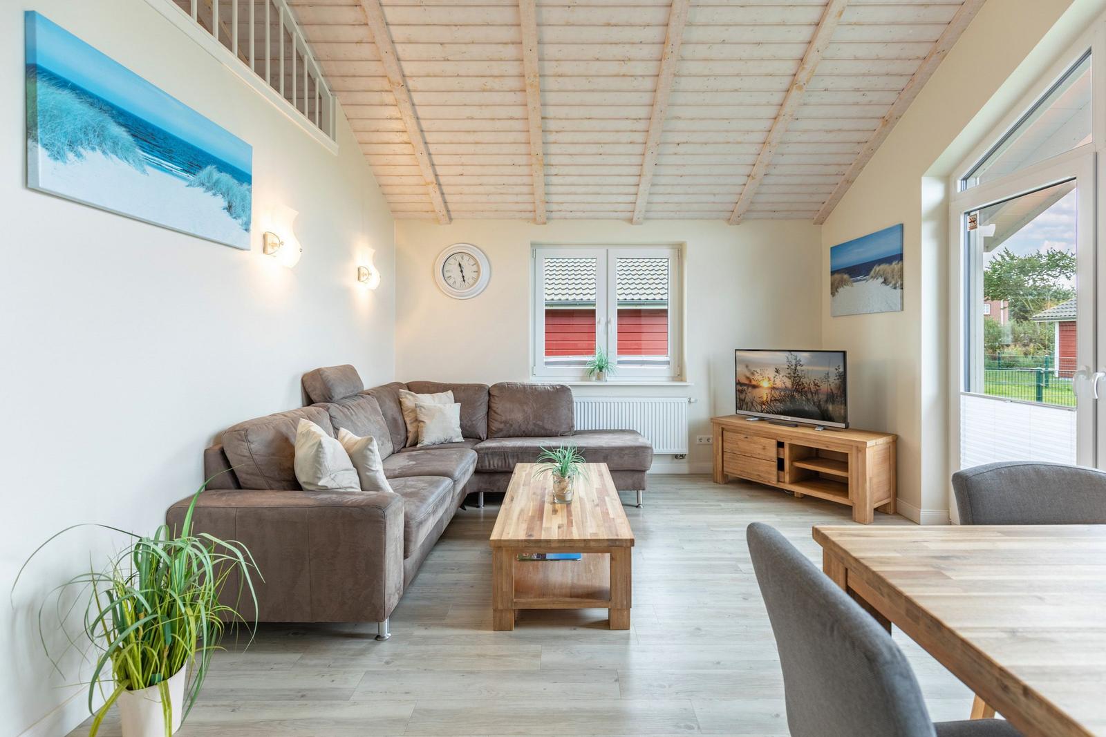 Wohnzimmer mit Sofa, Couchtisch und Fernseher. Holzdecke und große Fenster.