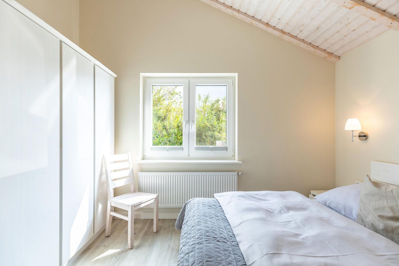Schlafzimmer mit Bett, Kleiderschrank, Stuhl und Fenster mit Blick auf Grün.