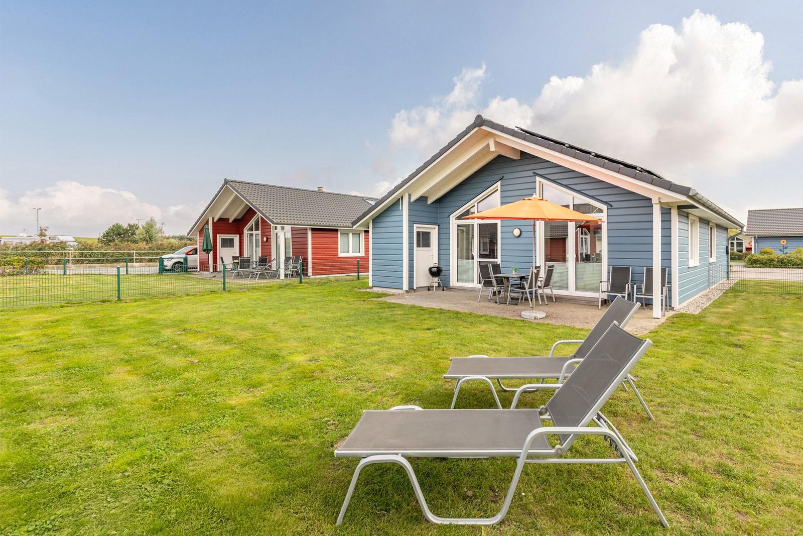 Blau gestrichenes Ferienhaus mit Garten, Sonnenliegen und Terrasse mit orangefarbenem Sonnenschirm.
