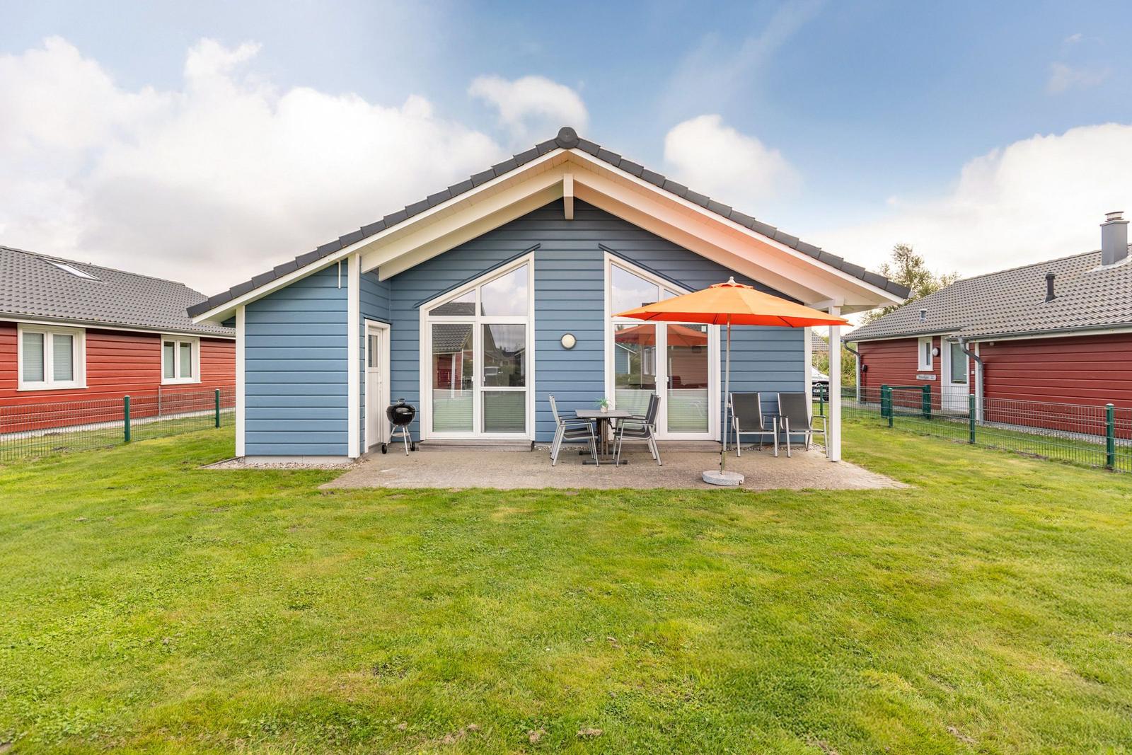 Blauer Ferienhaus mit Terrasse, orangefarbener Sonnenschirm und Grill.