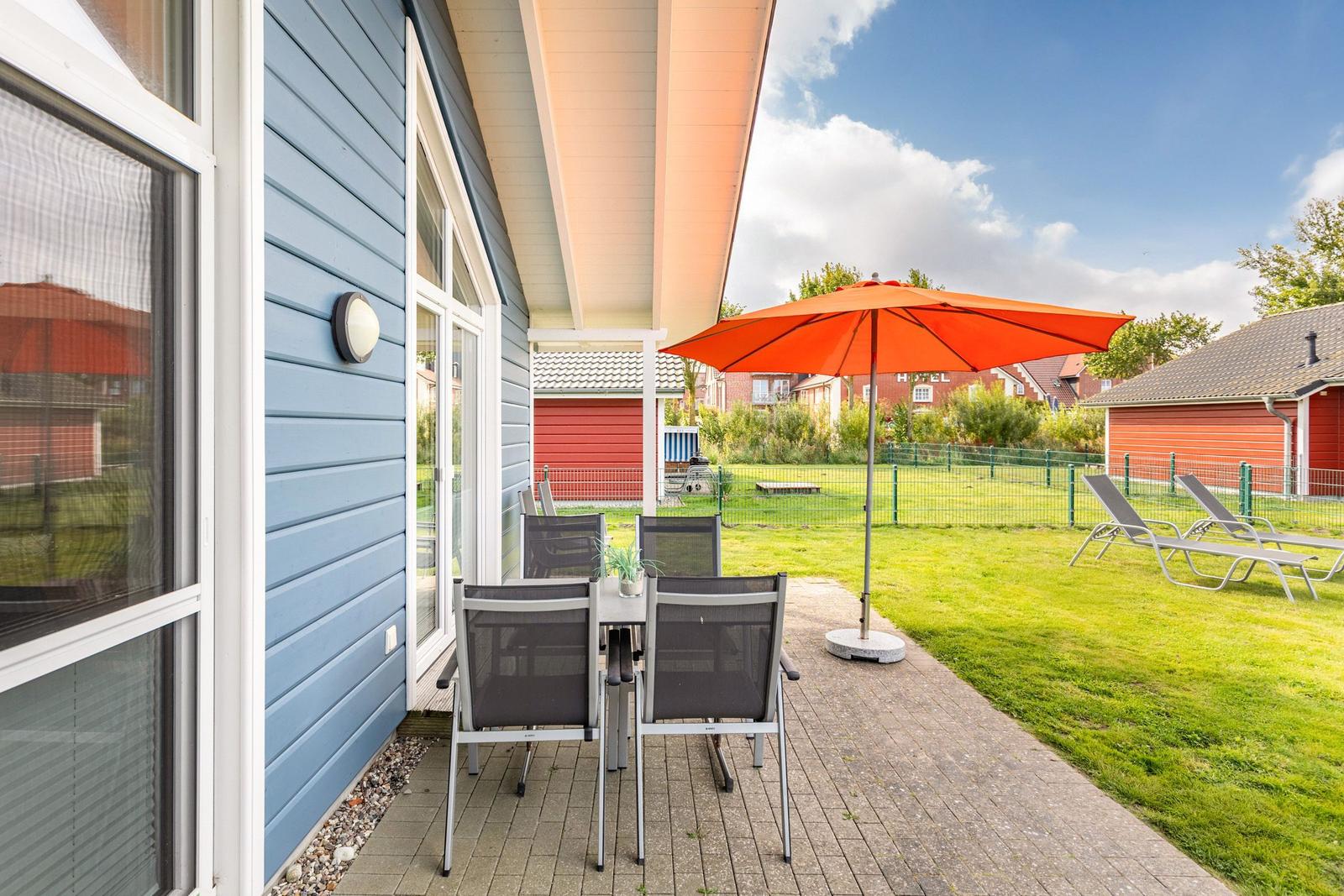 Terrasse mit Tisch, Stühlen und orangefarbenem Sonnenschirm. Grünfläche und Gartenmöbel im Hintergrund.