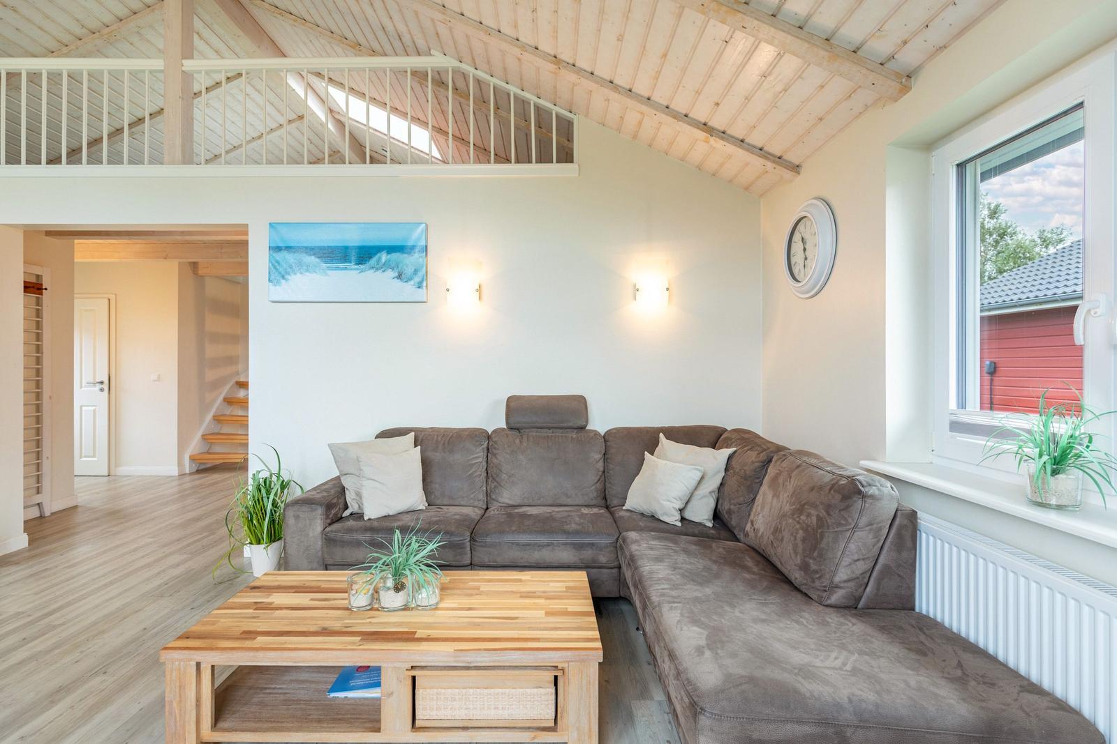 Wohnzimmer mit Ecksofa, Holztisch und Blick auf Balkon. Holzdecke und Wanduhr sichtbar.