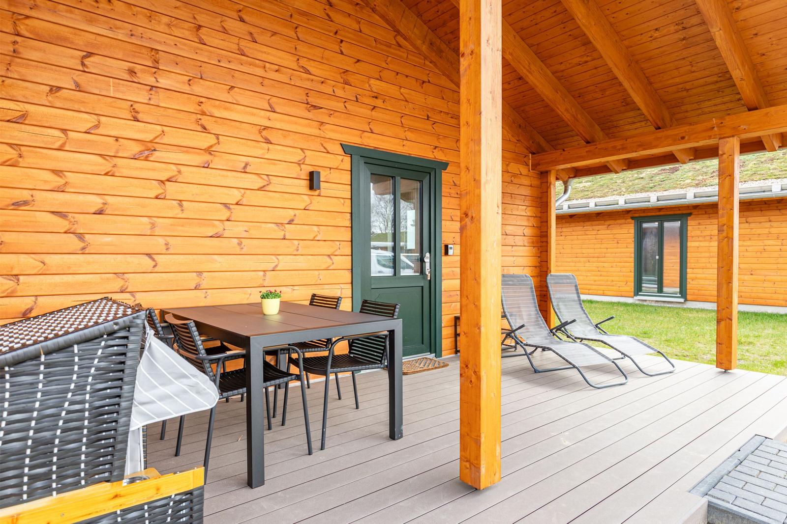 Terrasse mit Tisch, Stühlen und Liegestühlen an Holzhaus mit grüner Tür.