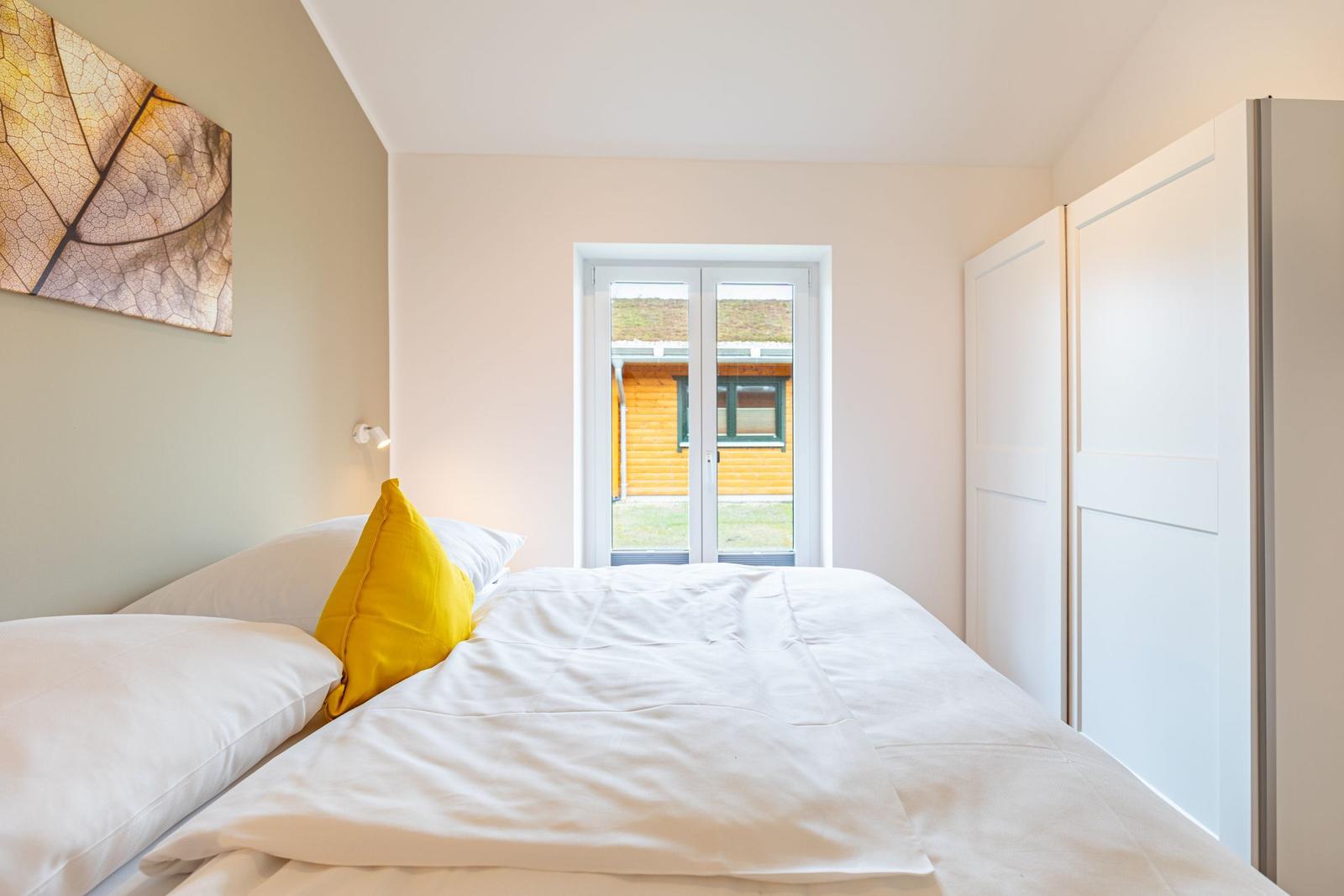 Schlafzimmer mit Bett, gelbem Kissen und Fensterblick auf Holzhaus.
