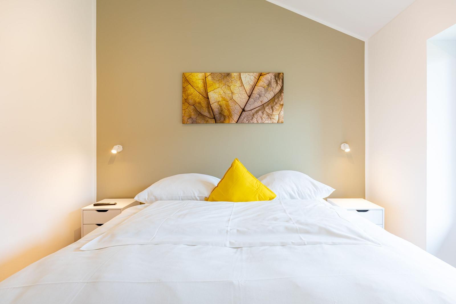 Schlafzimmer mit weißem Bett, gelbem Kissen und Wandbild von Blatt