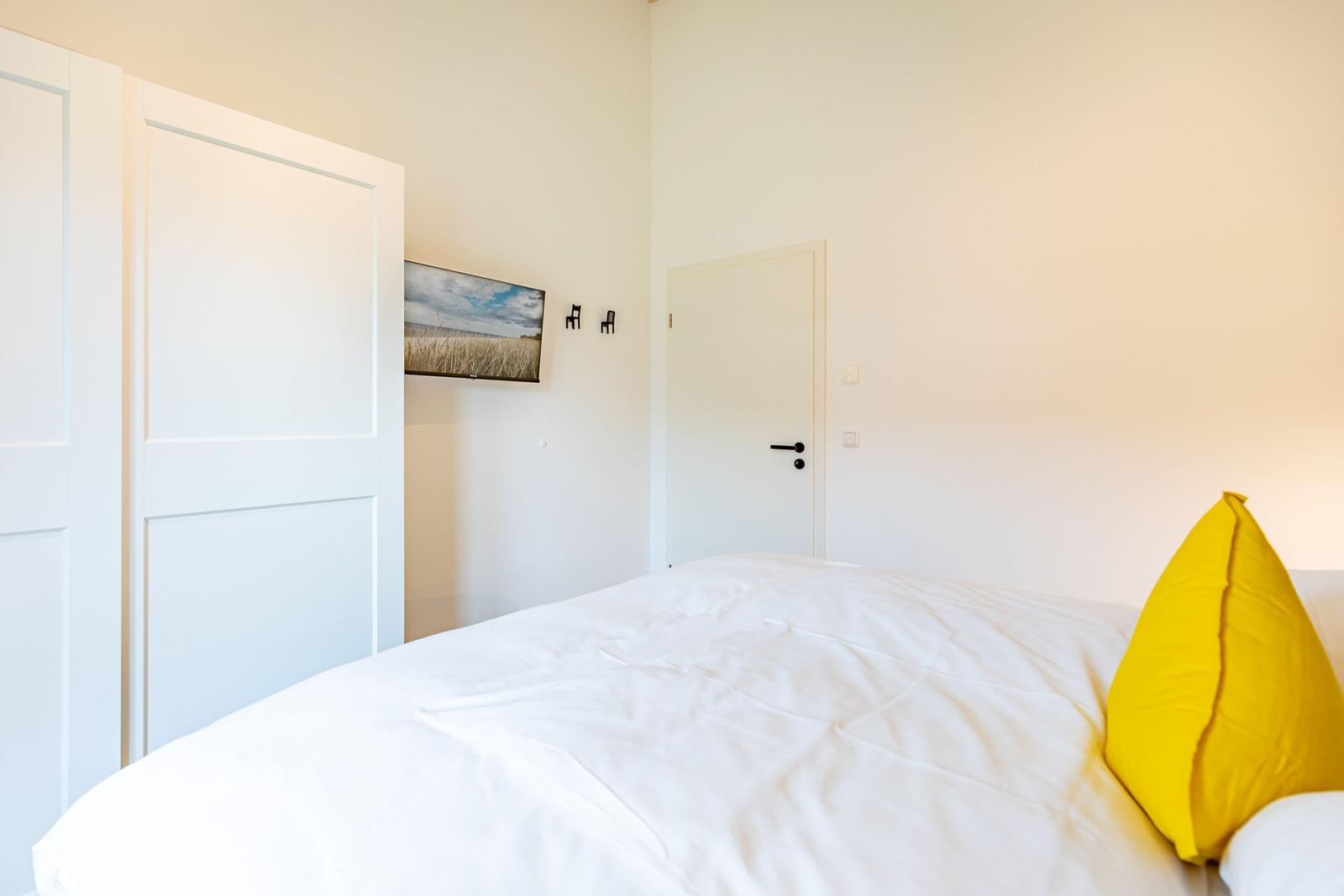 Schlafzimmer mit weißem Bett, gelbem Kissen, weißen Türen und Wandbild.