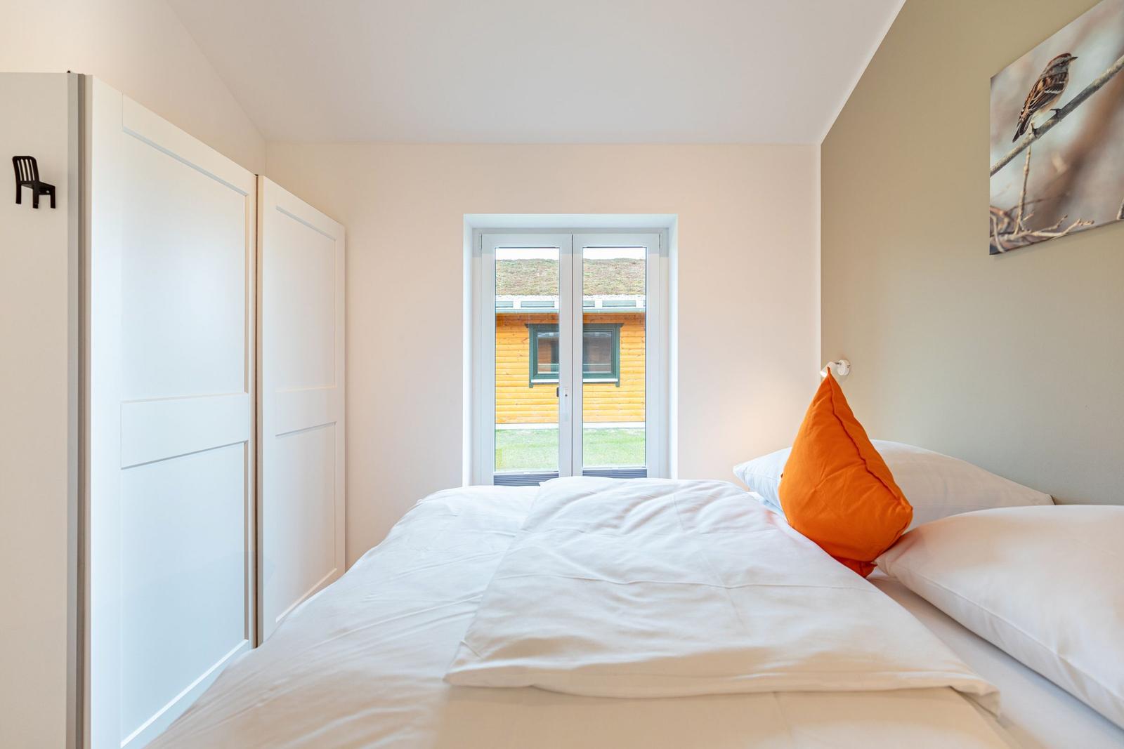 Schlafzimmer mit Bett, Fenster und Wandbild. Links Schrank, rechts Kissen.