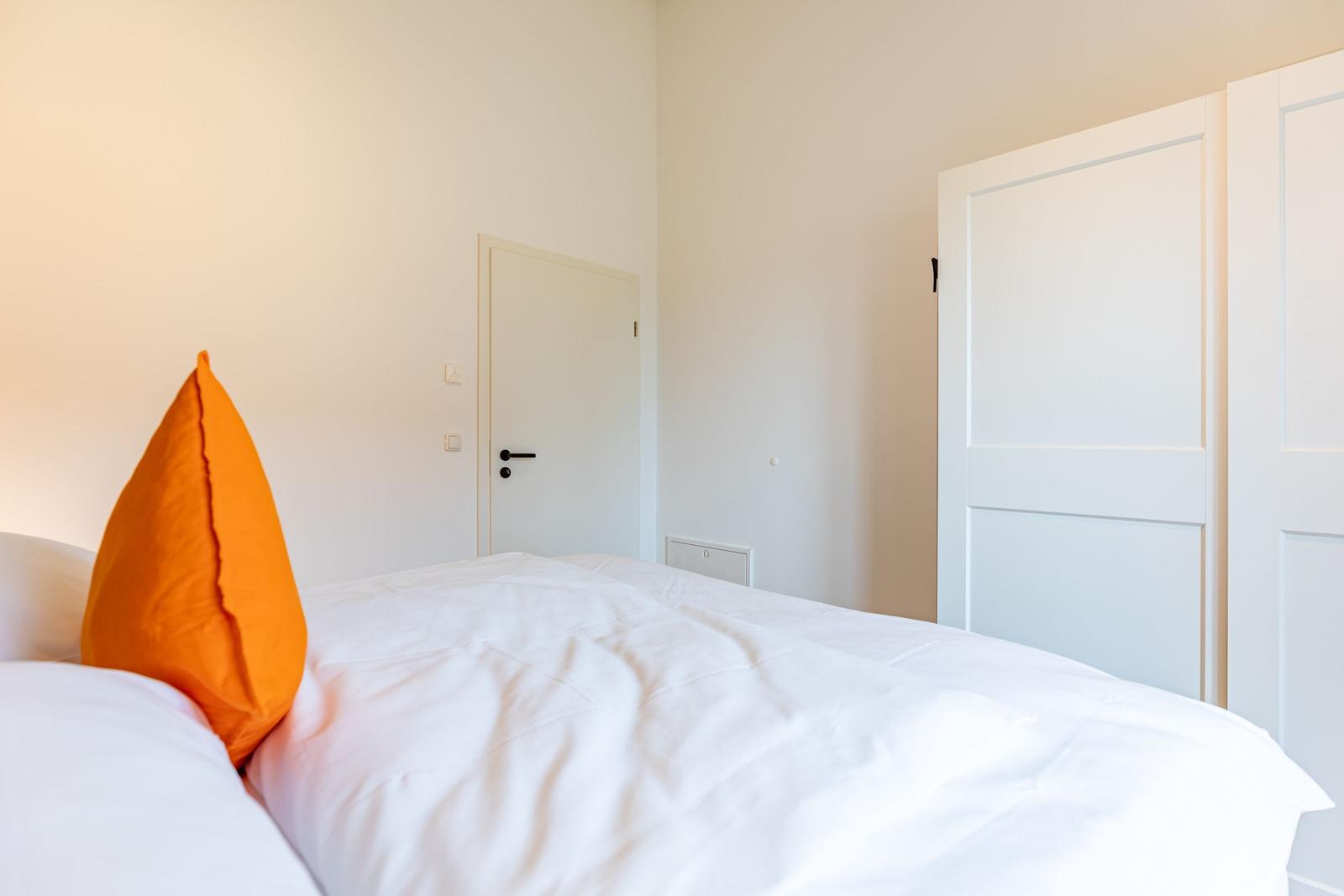 Schlafzimmer mit weißem Bett und orangefarbenem Kissen. Weiße Türen und Wände.