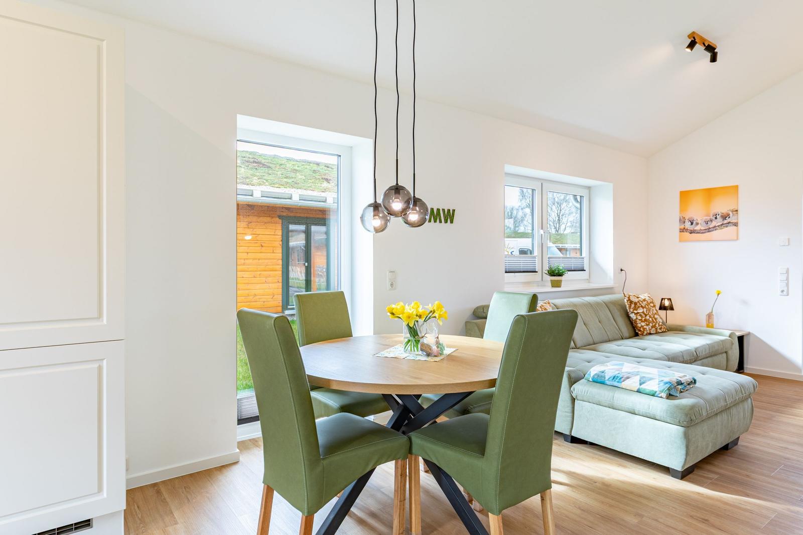 Essbereich mit runden Tisch und grünen Stühlen, Wohnbereich mit Sofa und Fenstern.
