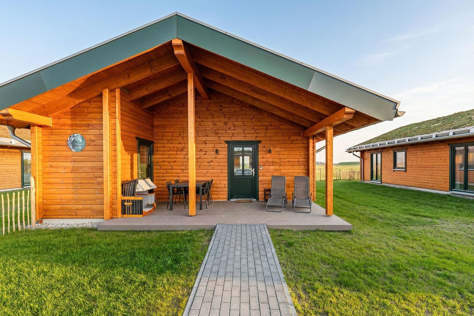 Holzhaus mit Terrasse, Tisch, Stühlen und Liegestühlen. Grasfläche und Pfad führen zum Eingang.