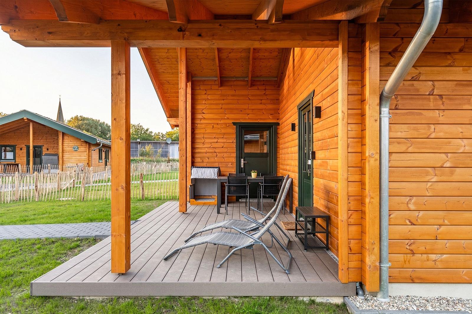 Terrasse mit Liegestühlen und Tisch unter Holzüberdachung. Hintergrund zeigt grünen Rasen und weitere Holzhäuser.