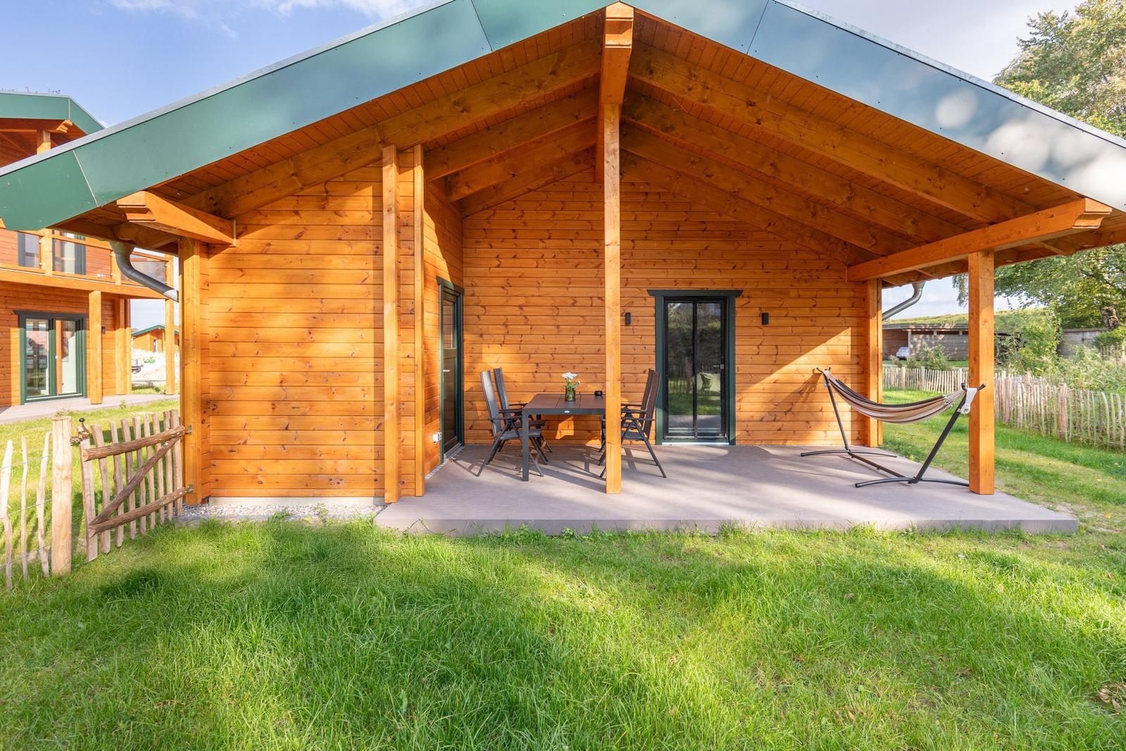 Holzhaus mit Terrasse, Tisch, Stühlen und Hängemöbel. Grasfläche und Zaun im Hintergrund.