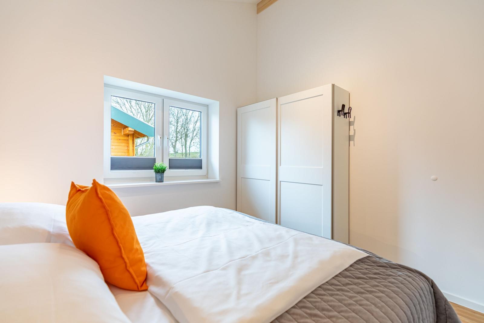 Schlafzimmer mit Bett, Fenster und Kleiderschrank. Draußen ist ein Holzhaus sichtbar.