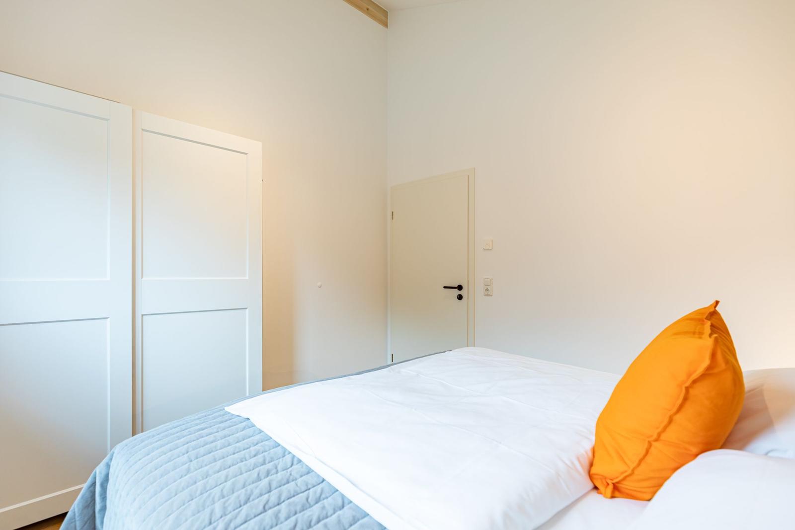 Schlafzimmer mit Bett, orangefarbenem Kissen und weißen Wänden.