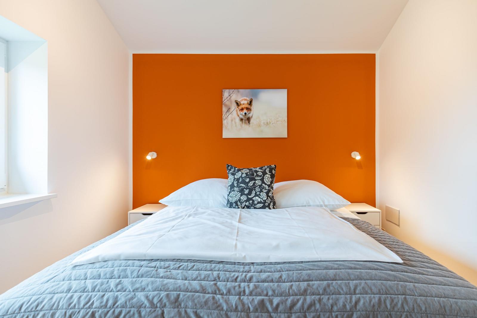 Schlafzimmer mit orangefarbener Wand, Bett mit weißer Bettwäsche und einem Kissen mit Muster.