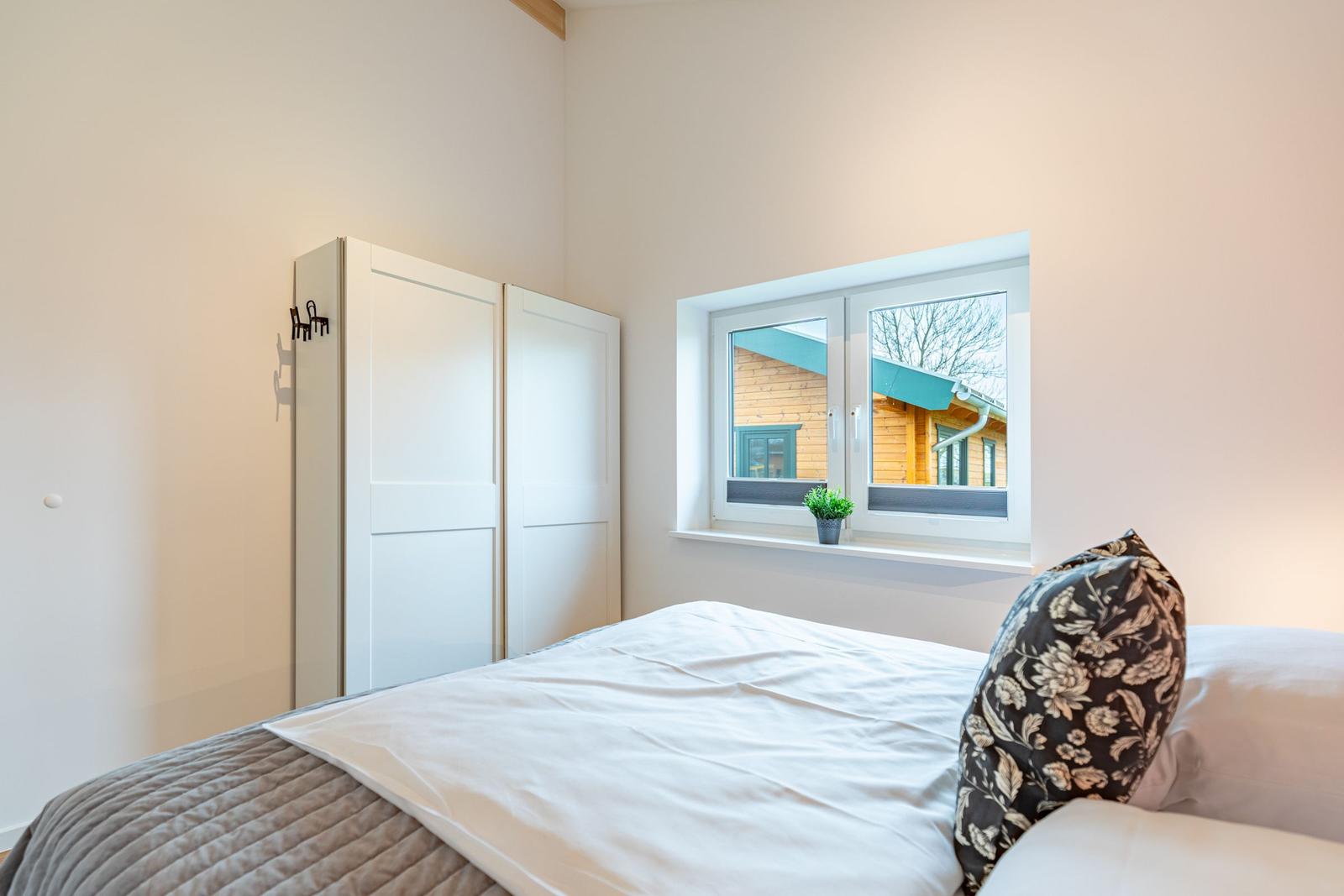 Schlafzimmer mit Bett, Kleiderschrank und Fensterblick auf Holzhaus.