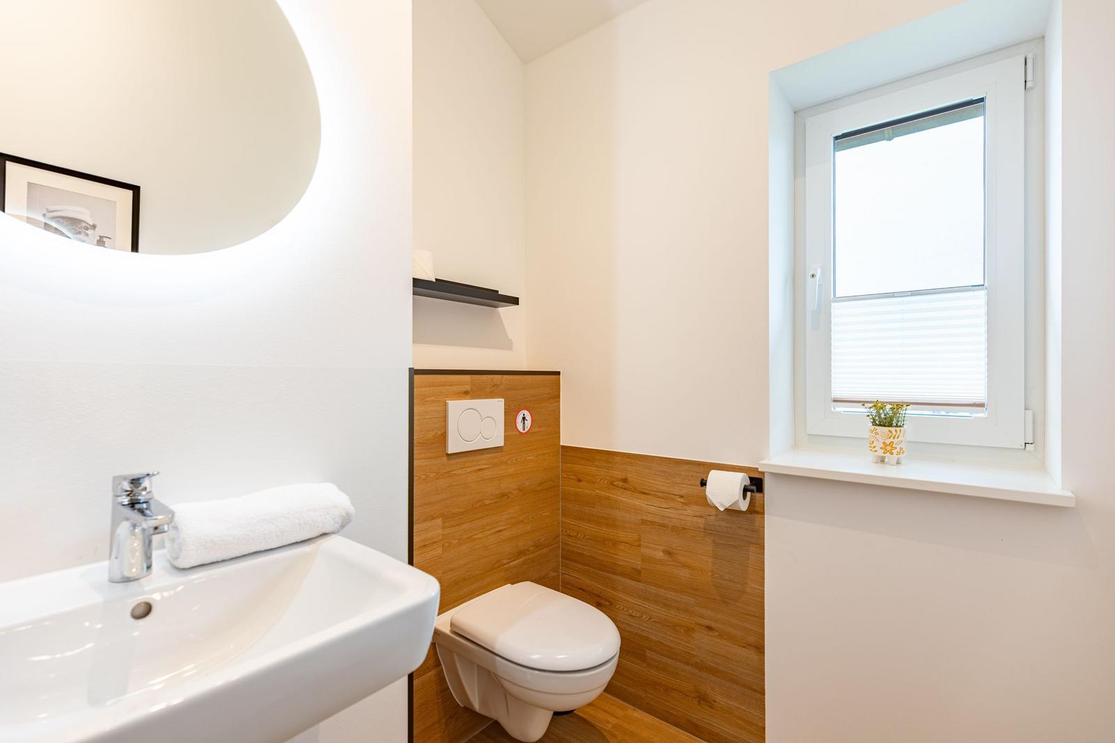 Badezimmer mit Waschbecken, Toilette und Fenster. Holzwand und Spiegel sichtbar.