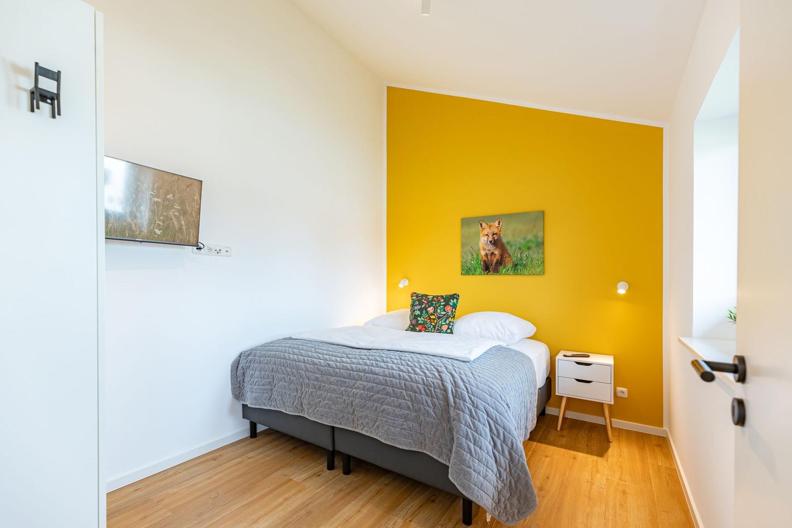 Schlafzimmer mit gelber Wand, Bett, Nachttisch und Fernseher an der Wand.