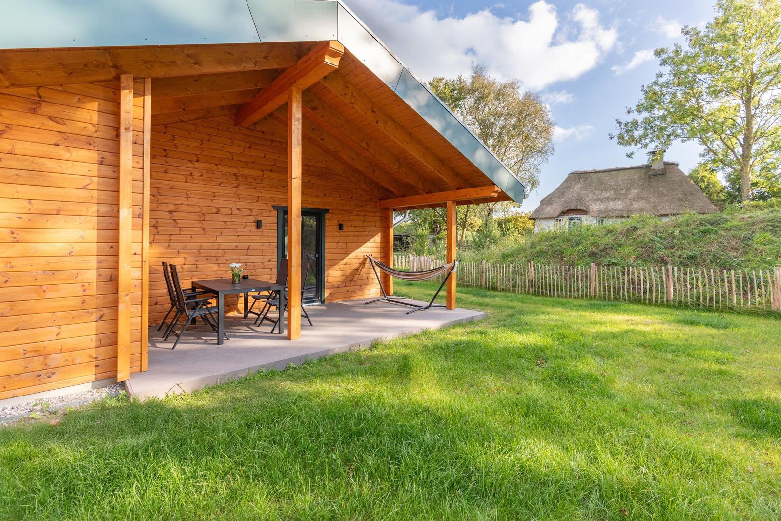 Holzhaus mit Terrasse, Tisch, Stühlen und Hängematte. Grasfläche und Zaun im Hintergrund.