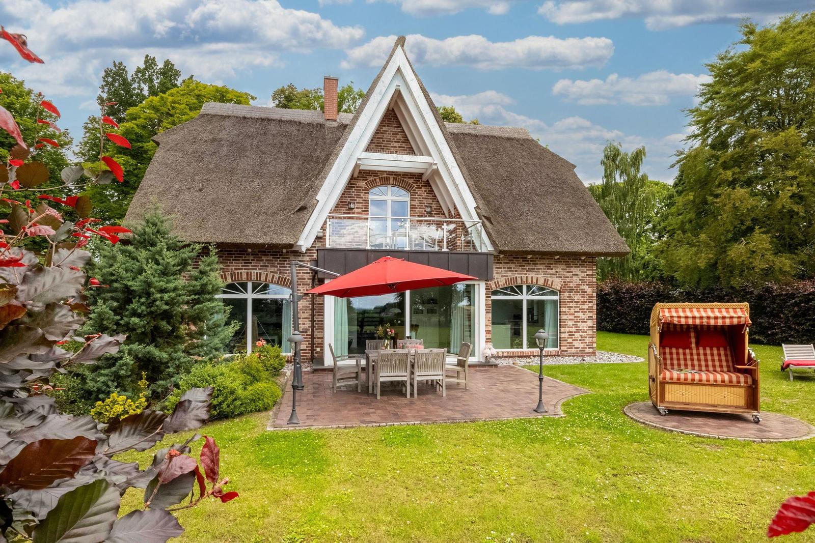 Haus mit Strohdach, Terrasse mit Tisch, Stühlen und rotem Sonnenschirm. Garten mit Liegestuhl und Bäumen.