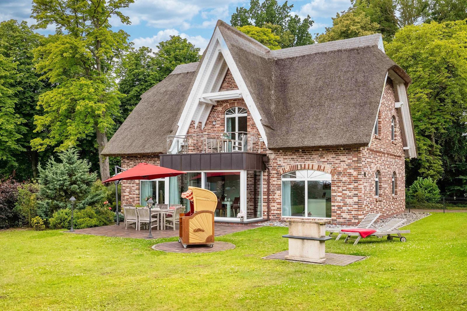 Ein Haus mit Strohdach, Terrasse und Garten mit Liegen und Sonnenschirm.