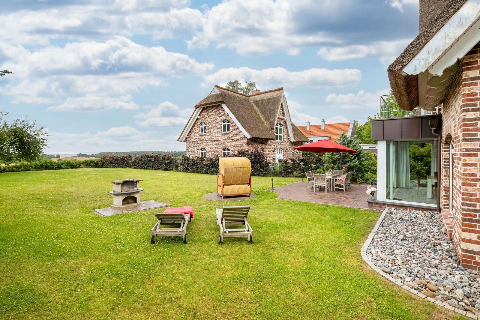 Haus mit Garten, Liegestühlen, Grill und Terrasse unter rotem Sonnenschirm.