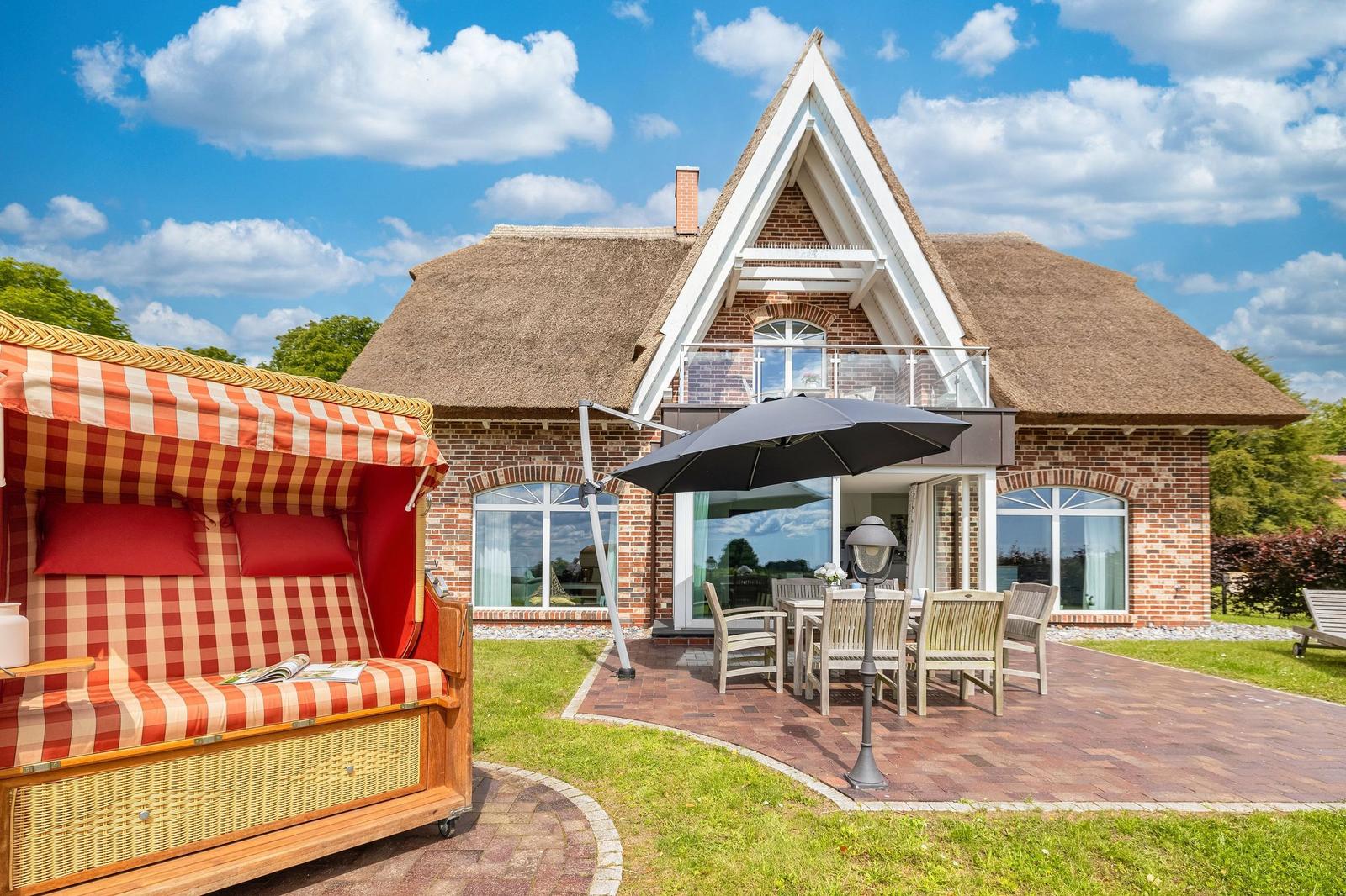 Haus mit Strohdach, Terrasse mit Tisch, Stühlen und Sonnenschirm. Links Strandkorb mit rot-weißem Muster.