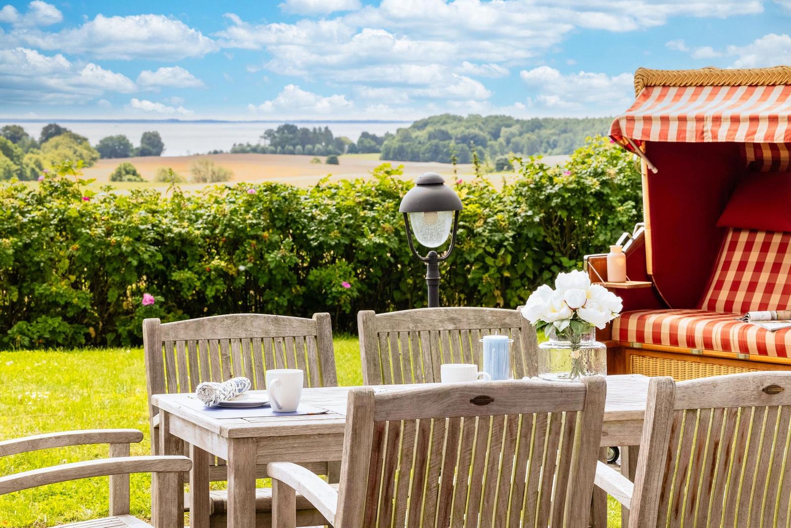 Terrasse mit Holztisch, Stühlen und Strandkorb. Blick auf Felder und Wasser.