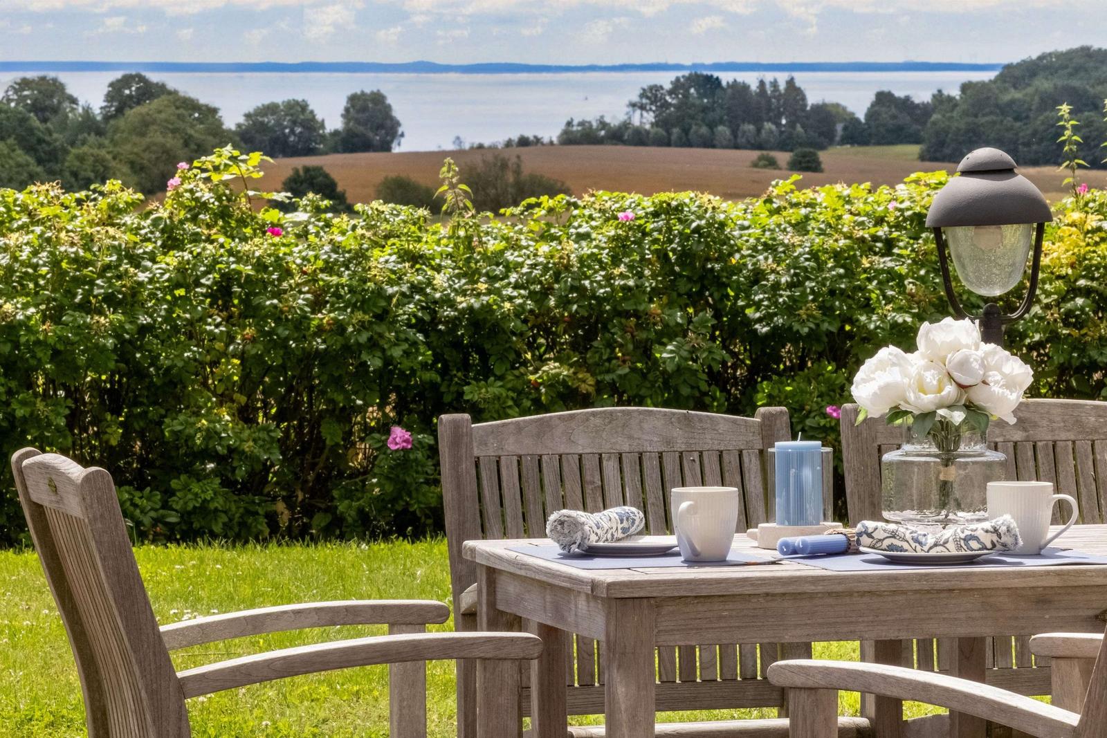 Holztisch mit Stühlen, Blumen und Kerzen auf einer Terrasse mit Blick auf Feld und Wasser.
