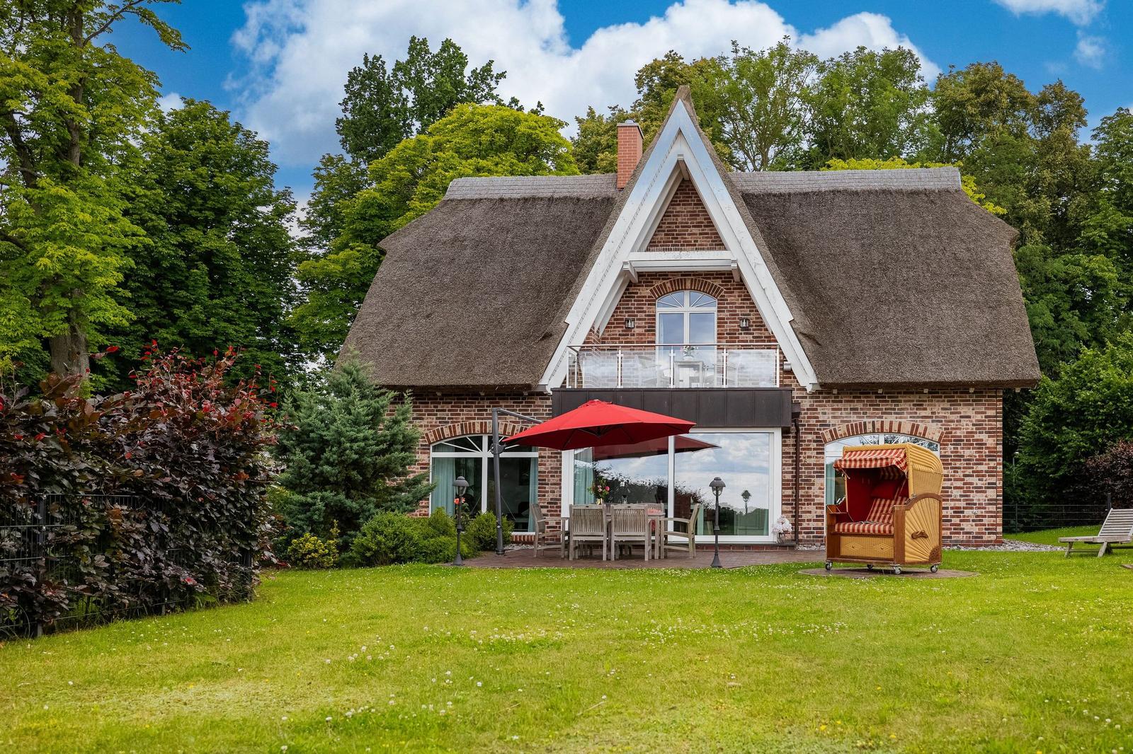 Haus mit Strohdach, Terrasse und Garten. Roter Sonnenschirm und Strandkörbe stehen draußen.