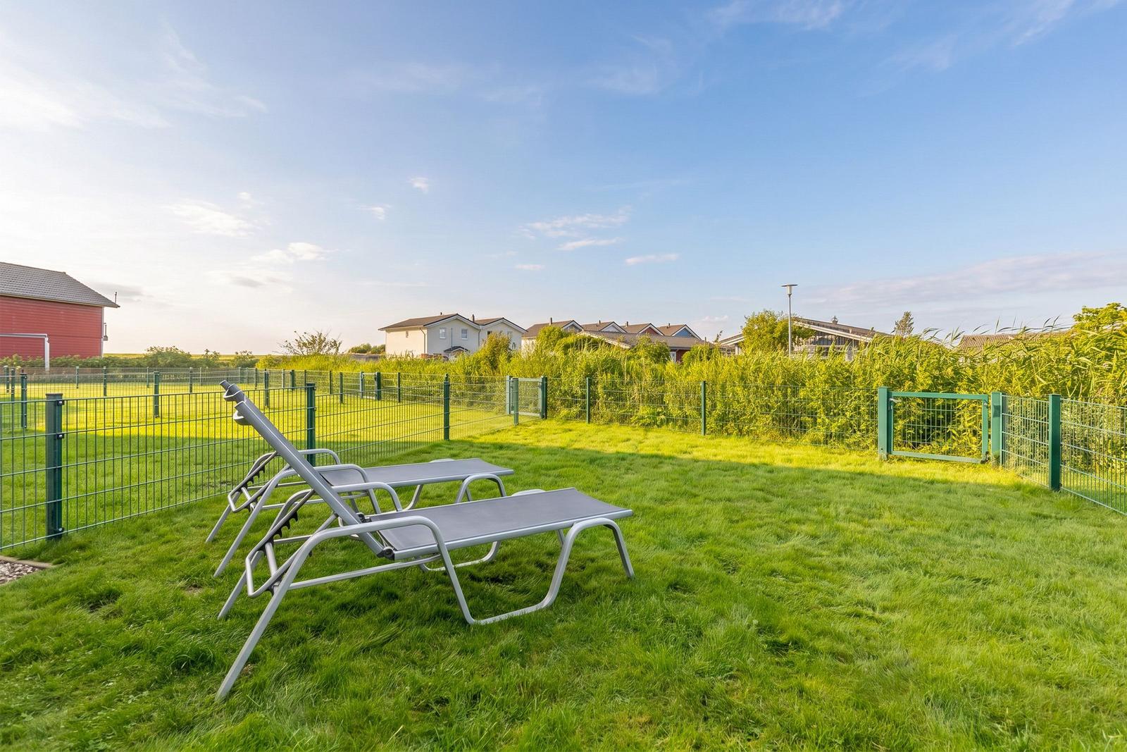 Two sun loungers on green grass in front of a fence with view of houses.