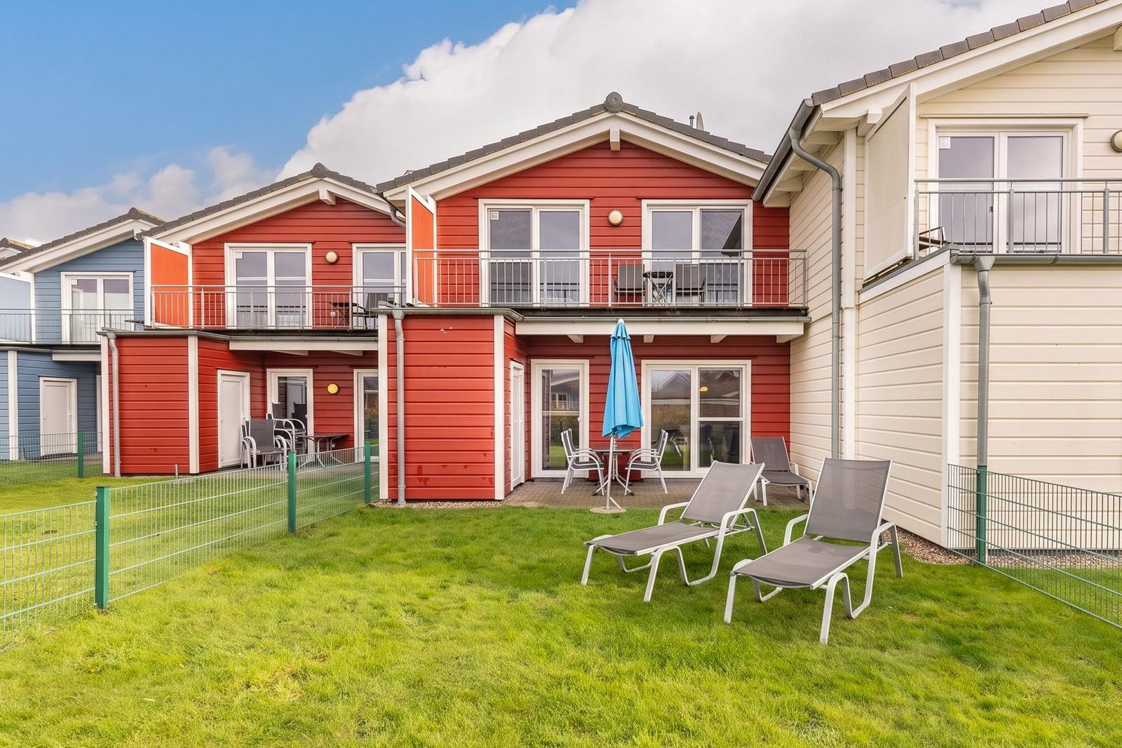 Red vacation home with balcony, garden, and sun loungers. Blue umbrella and terrace.