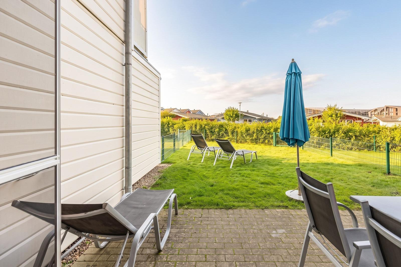 Terrace with lounge chairs and blue umbrella. Lawn and fence in the background.